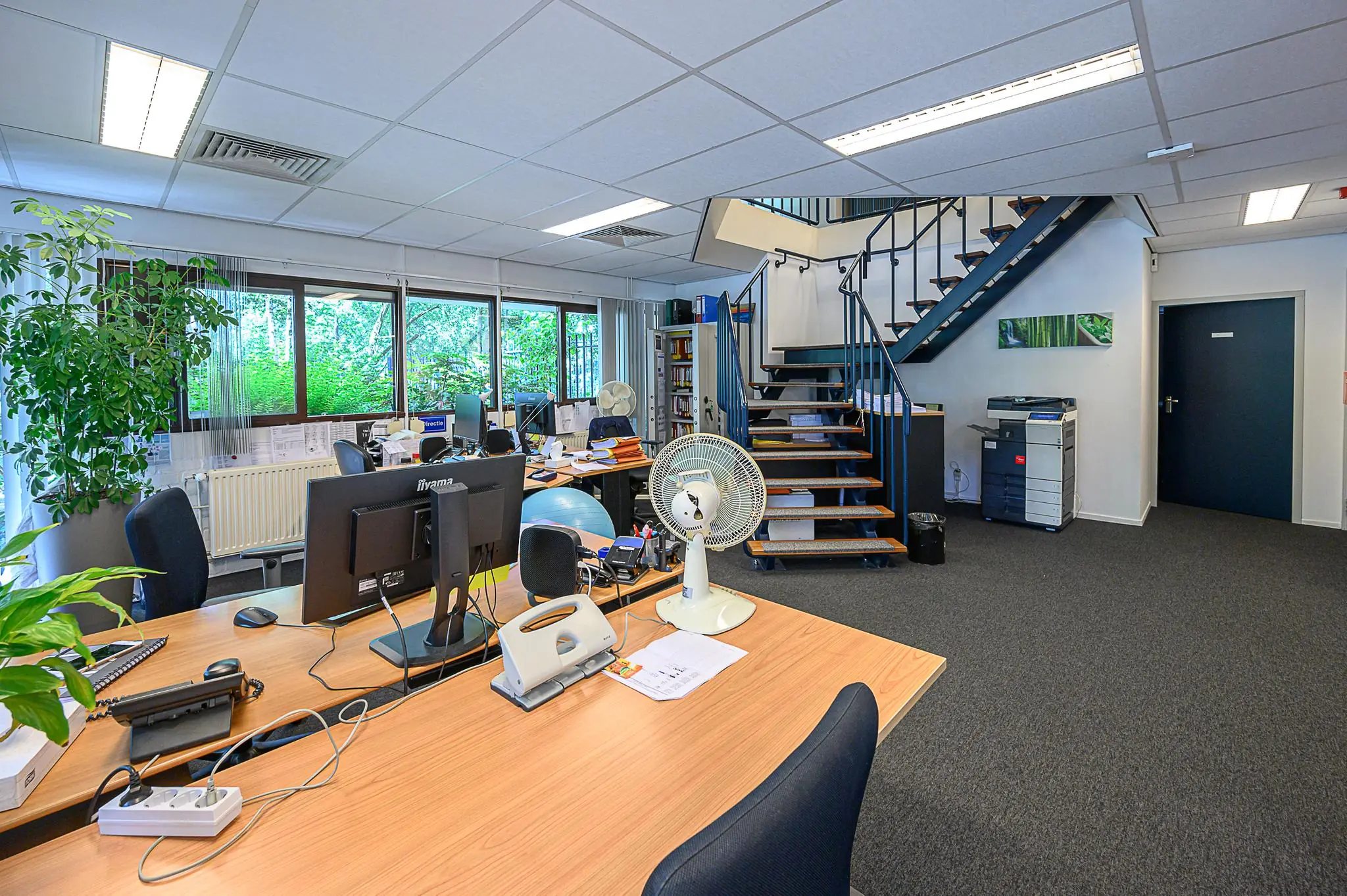 Modern kantoorinterieur met bureaus, computers, een trap en grote ramen met uitzicht op groen.